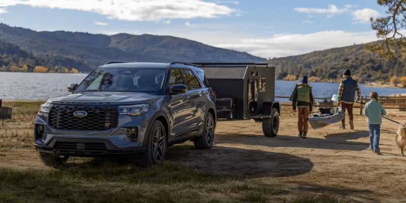 Gray Ford Explorer towing a camping trailer at a lakeside site with a family and dog nearby