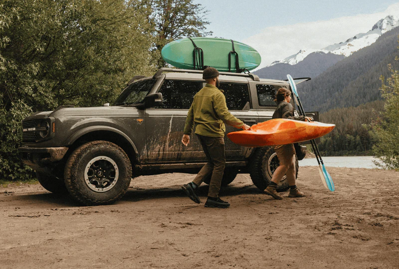 Two people carrying an orange kayak beside a muddy Ford Bronco with a surfboard on the roof rack
