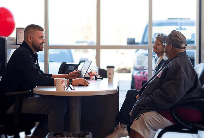 salesman and two buyers sitting on a desk in a dealership