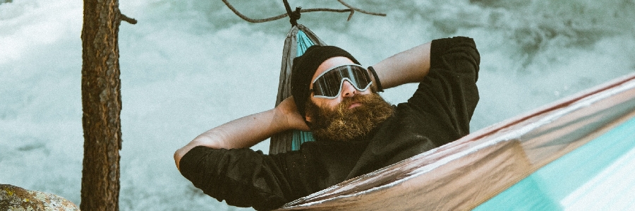 a BC man wearing sunglasses and a hat relaxes in a hammock above a river