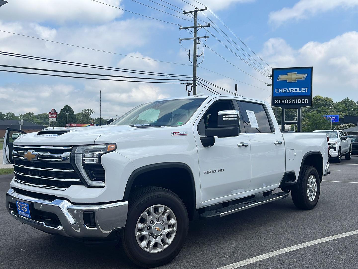 A white 2500HD pre-owned Silverado Z71 pickup truck in the front lot of Strosnider Chevrolet in a blue sky with summer puffy clouds.