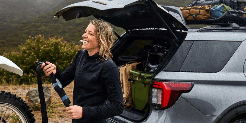 Smiling woman unloading gear from the trunk of a gray SUV on a rainy outdoor adventure