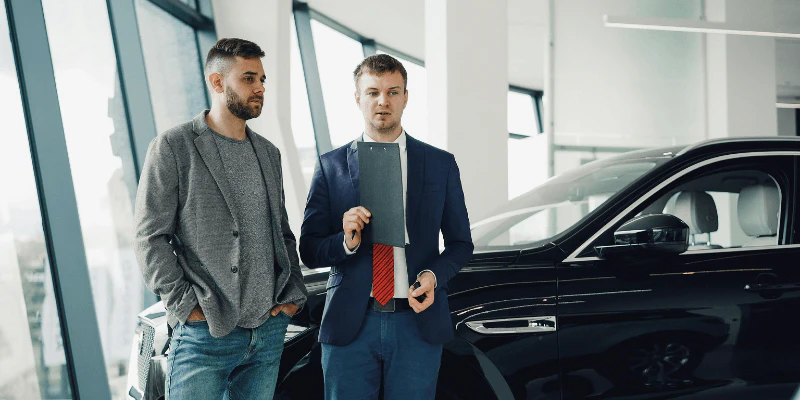 Car salesperson holding a clipboard speaking with a customer inside a dealership showroom