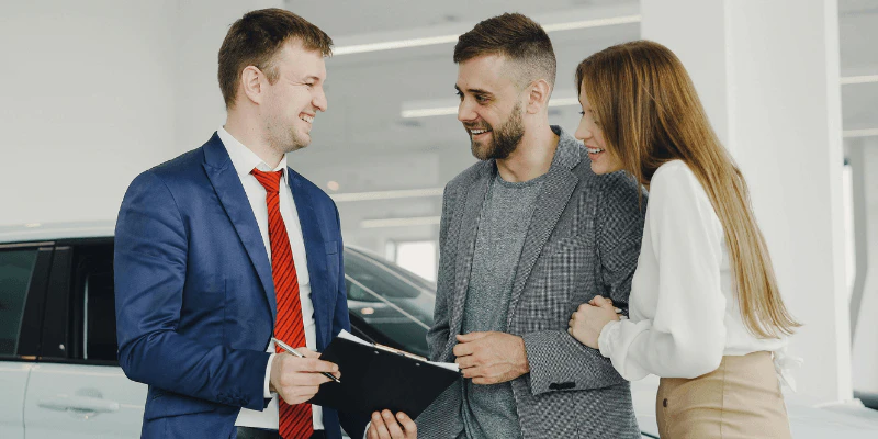 Smiling salesperson reviewing paperwork on a clipboard with a couple at a car dealership