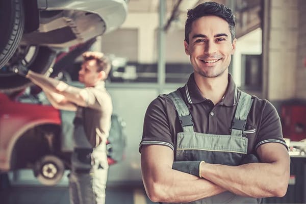 smiling service technician standing in front of a vehicle as a second technician works under the hood